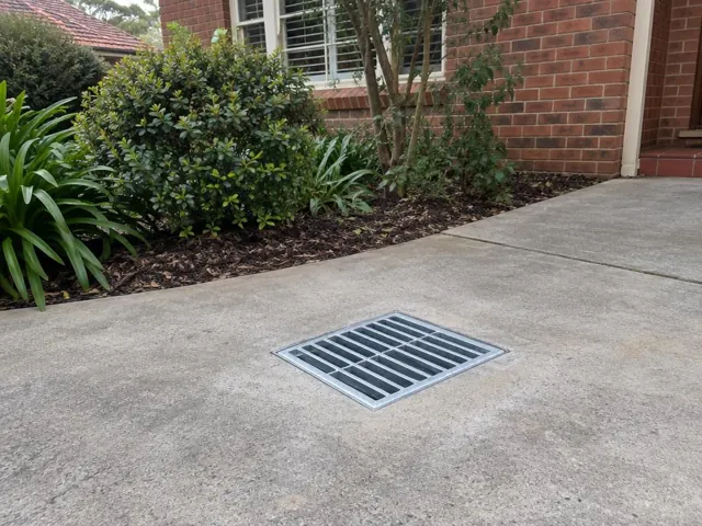 Cleared stormwater grate set in a concrete driveway beside brick house and garden beds in Pennant Hills